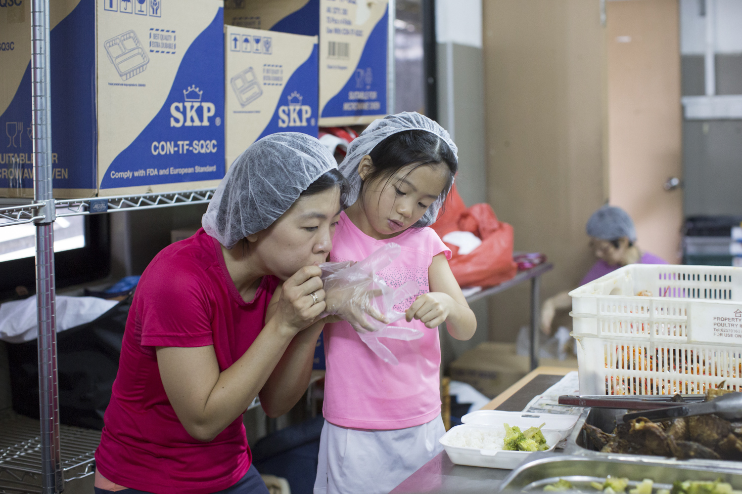 Dorothy helps Christine’s younger sister, Angie, put on plastic gloves as they help with packing meals at Willing Hearts.